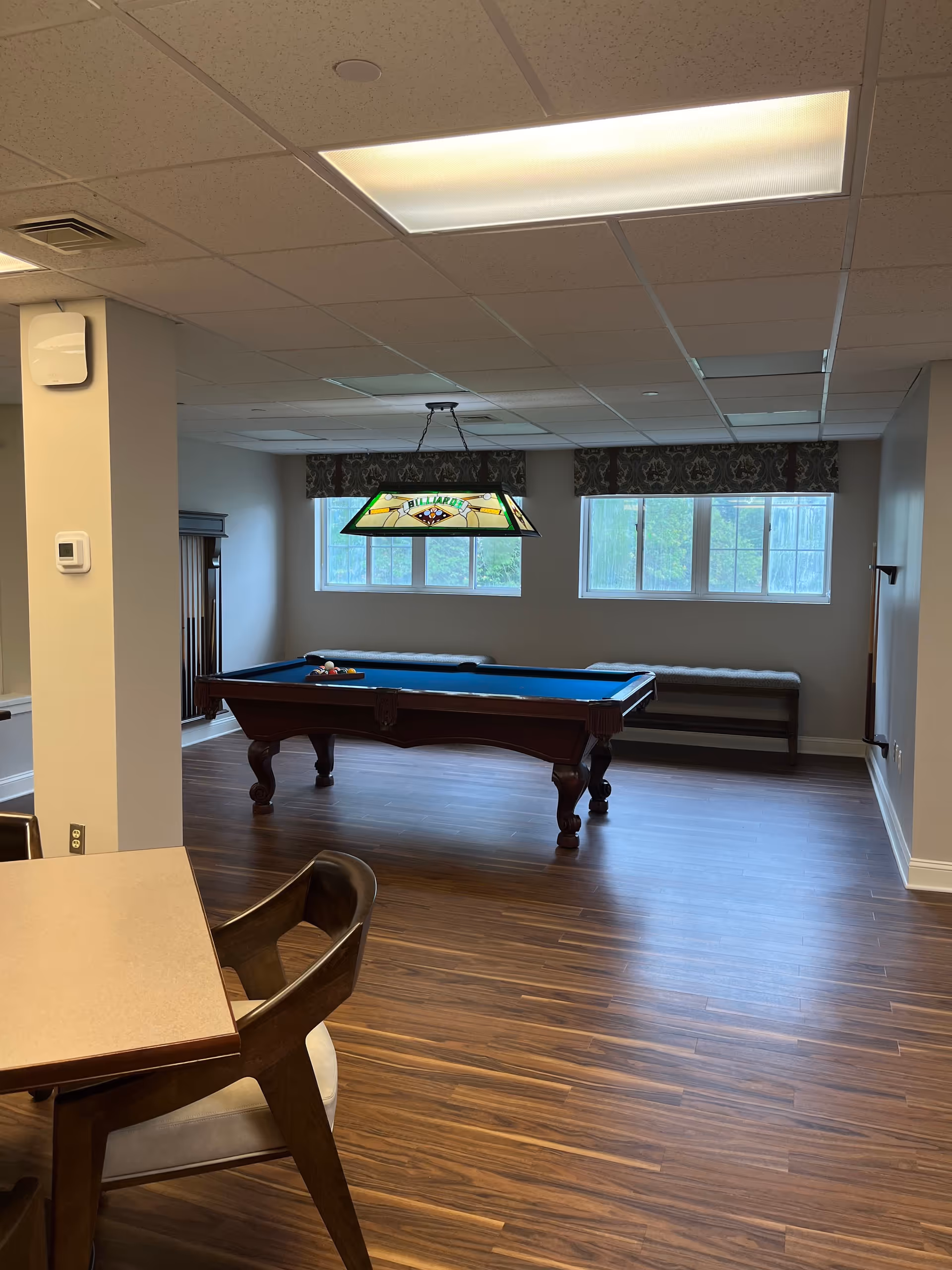 Interior room with a pool table in the center, a stained glass billiards light fixture hanging above it, two windows with patterned valances, a bench along the wall, and a table with chairs in the foreground. The room has wood flooring and a drop ceiling with fluorescent lighting.