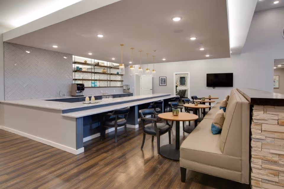 A modern dining area with a long marble countertop bar featuring black chairs, pendant lights hanging above, and small round wooden tables with chairs and a cushioned bench along the wall. The space has wood flooring, a wall-mounted TV, and shelves with glassware and decorative items.