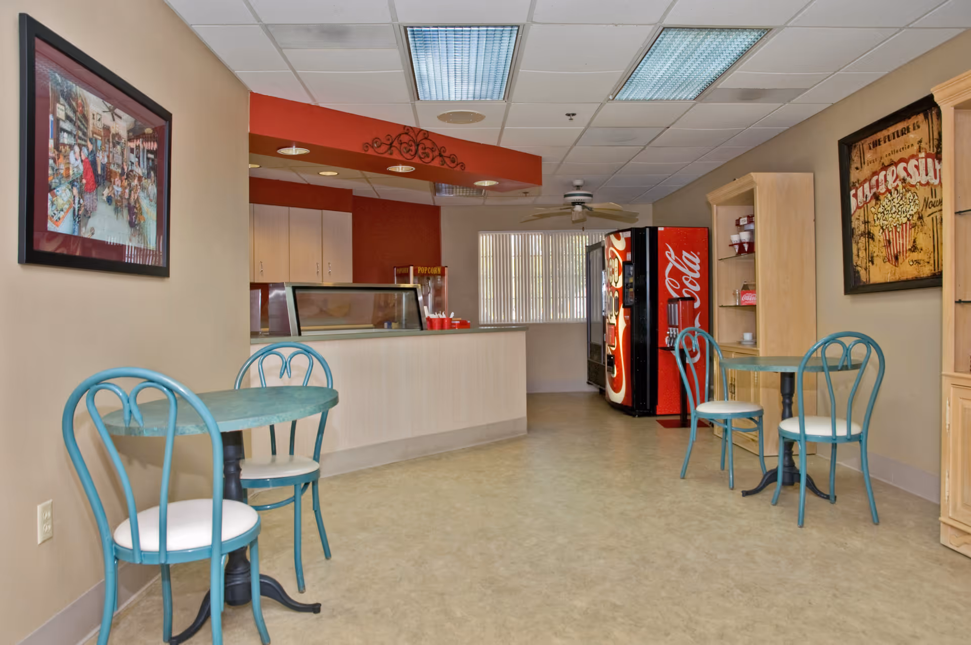 A small dining area with two round tables and four teal metal chairs with white cushions. In the background, there is a counter with a popcorn machine and a Coca-Cola vending machine. The walls are beige with framed pictures, and there is a ceiling fan and fluorescent lighting overhead.