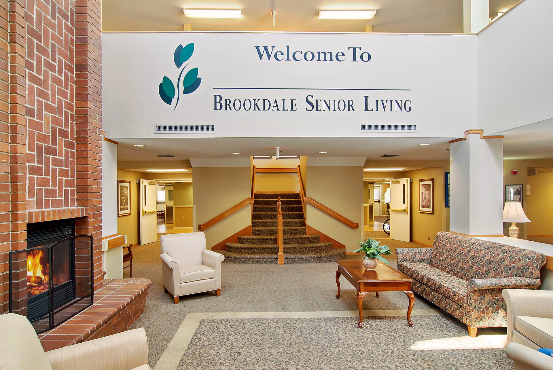 Interior view of a senior living facility lobby with a brick fireplace on the left, a beige armchair, a floral-patterned sofa, a wooden coffee table with a plant, and a carpeted staircase in the center. Above the staircase is a sign that reads 'Welcome To Brookdale Senior Living'.