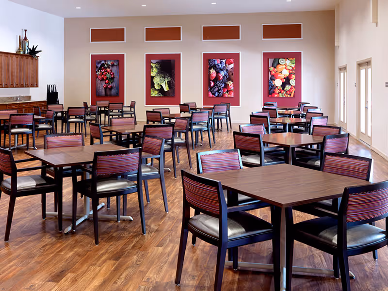 A spacious dining room with multiple wooden tables and chairs arranged neatly on a wooden floor. The walls are decorated with large framed pictures of various fruits and vegetables. There are windows and doors on the right side allowing natural light to enter the room.