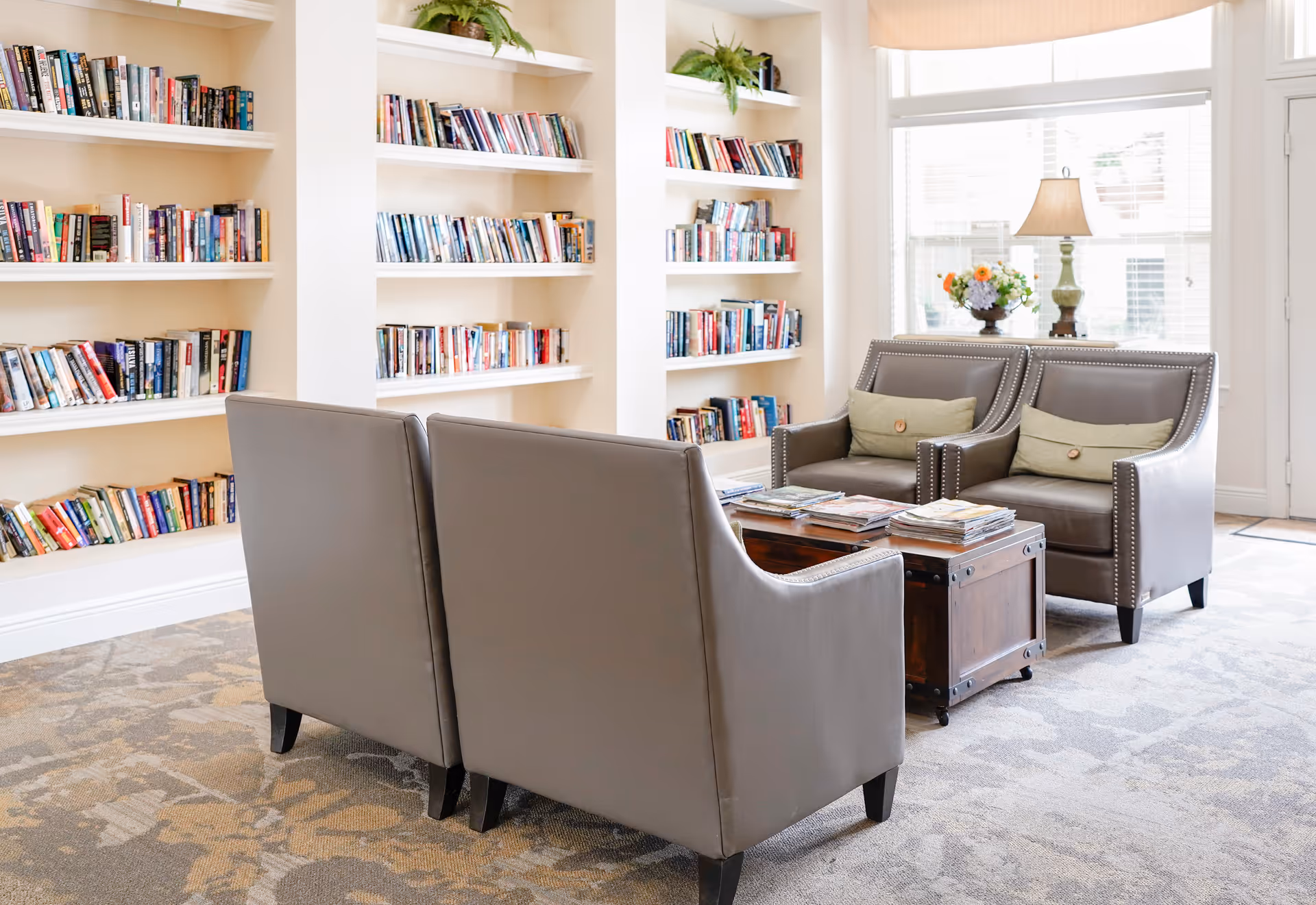 A cozy reading area in a senior living facility featuring built-in white bookshelves filled with books, two gray armchairs with green cushions, a wooden coffee table with magazines, a table lamp, and a window with blinds letting in natural light.