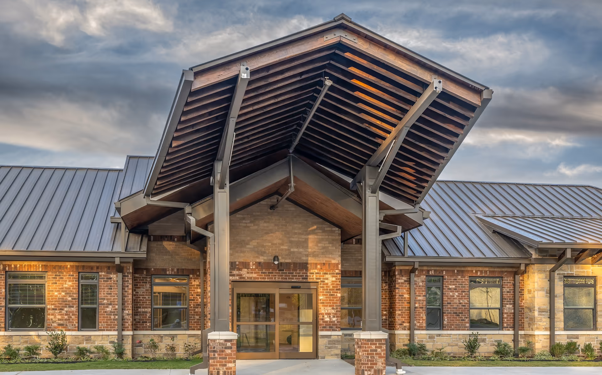 Front exterior view of a senior living facility with a large covered entrance supported by metal beams, brick and stone walls, and a metal roof under a cloudy sky.