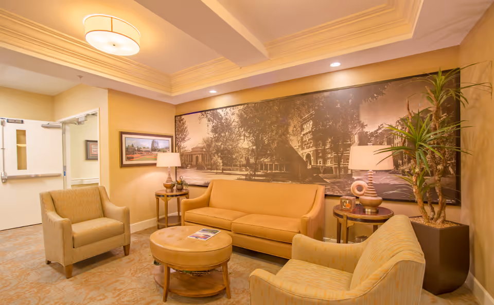 A cozy seating area with a sofa, two armchairs, a round ottoman, side tables and lamps beneath a large framed mural in a warmly lit common room.