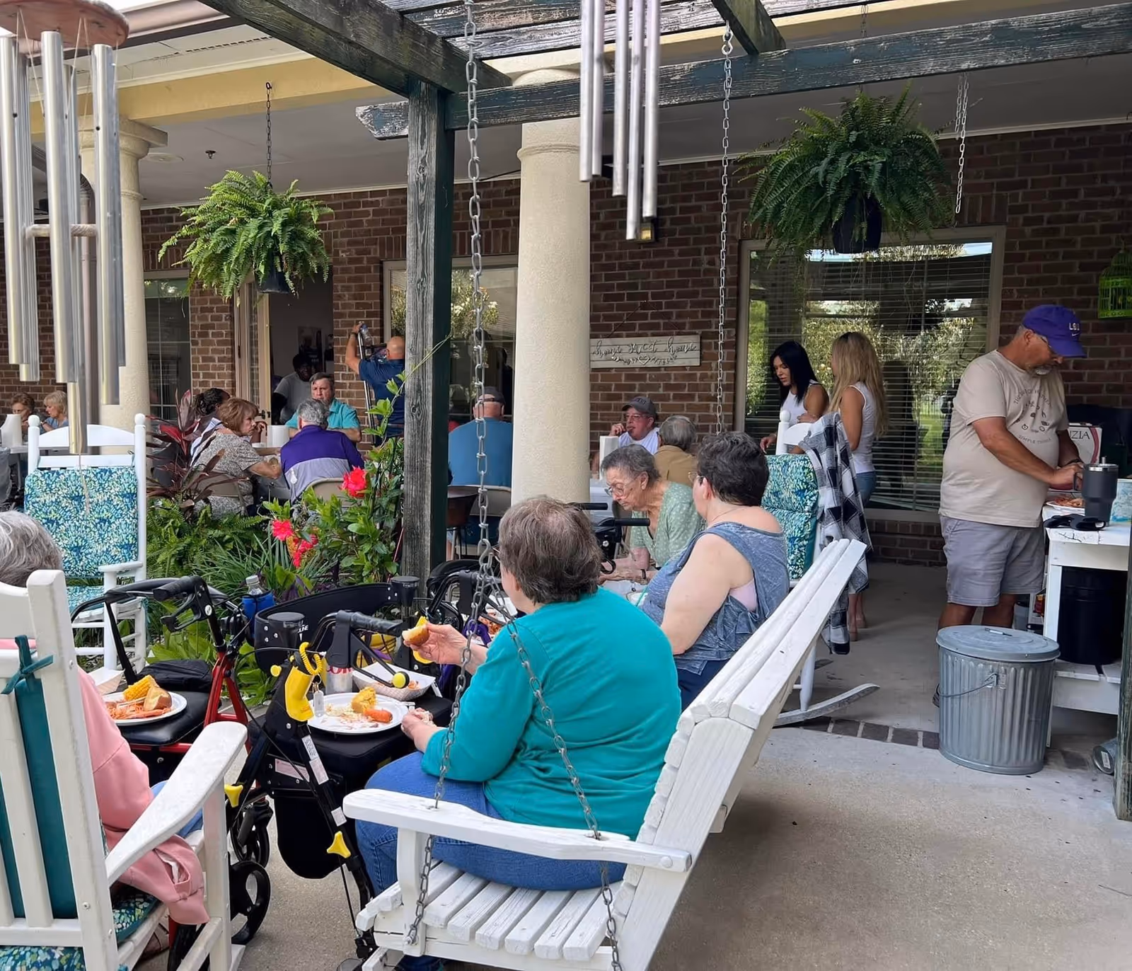 A group of elderly people and caregivers gathered in an outdoor covered patio area at a senior living facility. Several seniors are seated on white wooden swings and chairs, eating food from plates. There are hanging plants and wind chimes decorating the space, with a brick wall and windows in the background. A man in a purple cap is preparing food at a table on the right side.