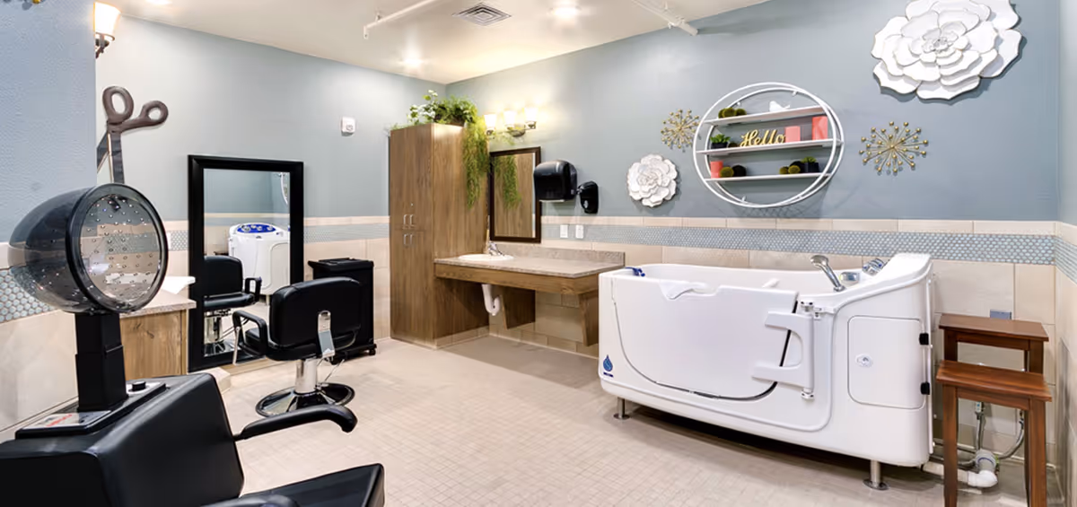 Interior room with salon chairs, a hooded hair dryer, mirrors and a walk-in bathtub against a blue-gray wall with decorative shelving.