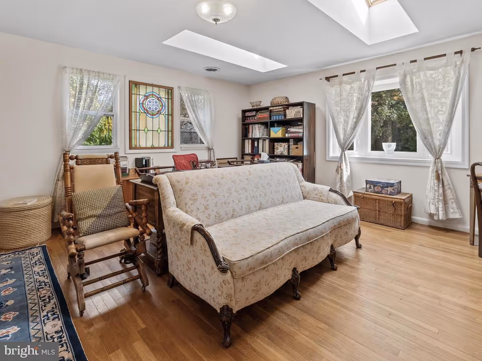Sunlit living room with a floral upholstered sofa, wooden rocking chair, bookshelf, and curtained windows.