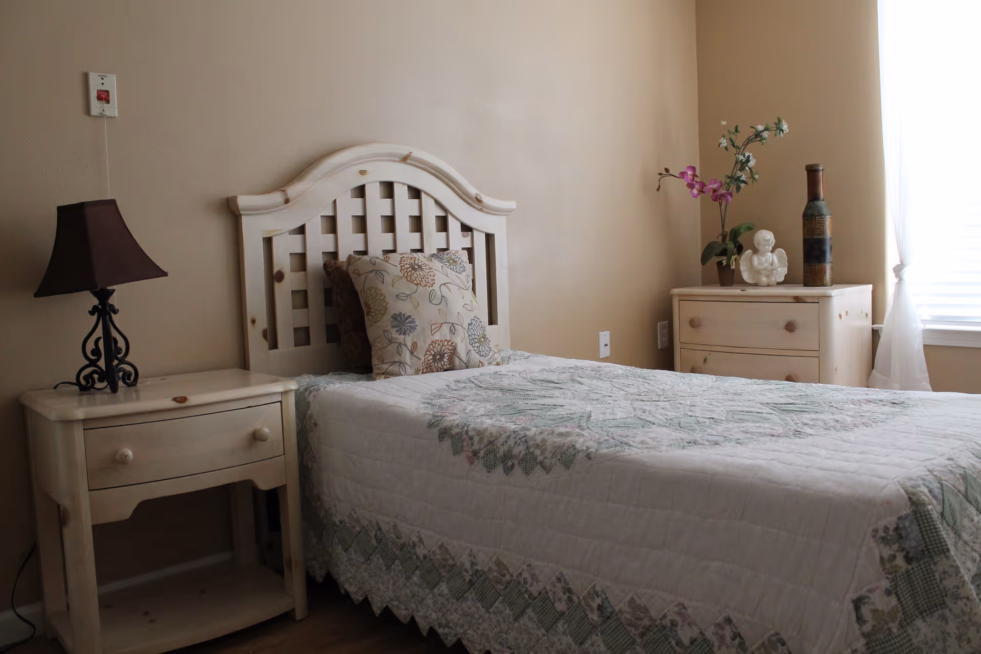 A neatly made single bed with a patterned quilt, headboard, two nightstands, a lamp and a bright window in a beige bedroom.