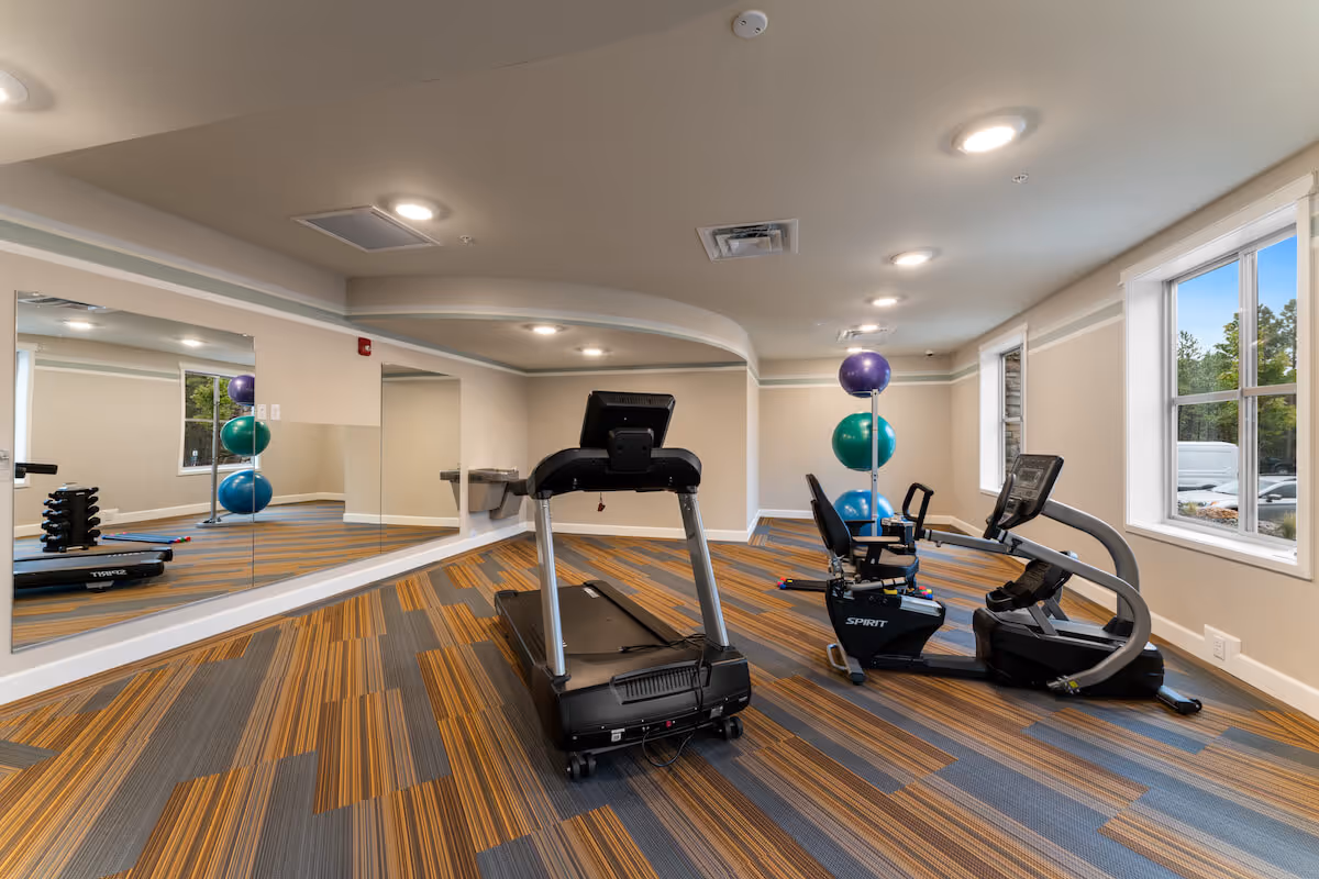 A fitness room in a senior living facility featuring a treadmill and a recumbent exercise bike on a carpeted floor with orange and gray stripes. There are large mirrors on one wall, three exercise balls stacked on a stand, and two windows letting in natural light. The room has beige walls and ceiling lights.
