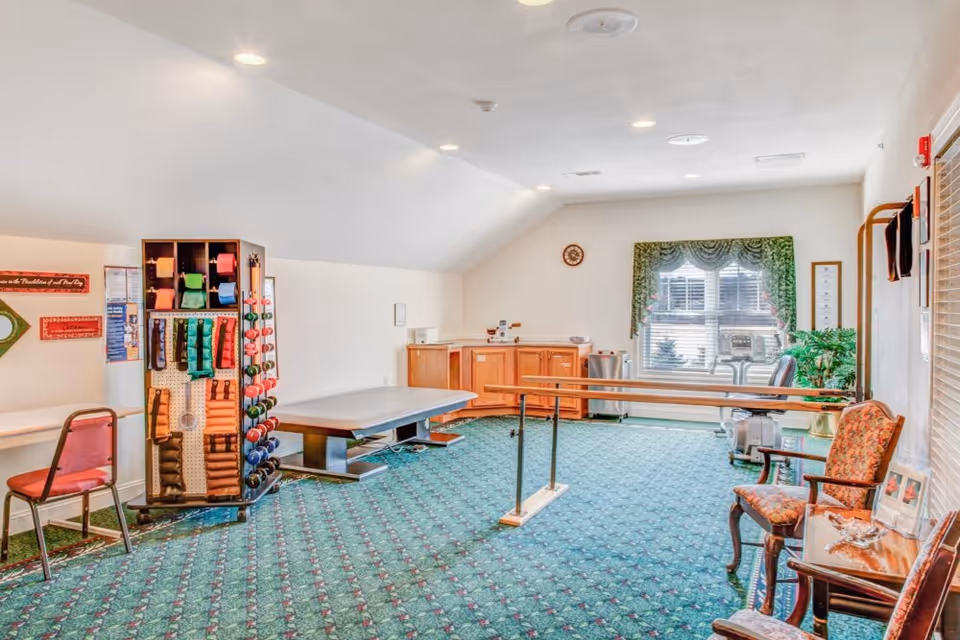 Bright therapy/exercise room with parallel bars, treatment table, weights rack, and seating.