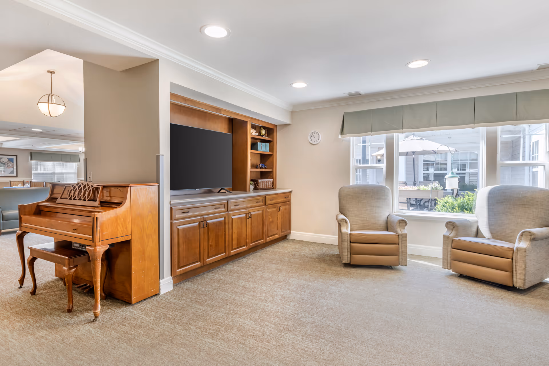A bright living room area in a senior living facility with two beige and brown armchairs positioned near large windows with a view of an outdoor patio. There is a wooden piano with a matching bench on the left side, and a built-in wooden cabinet with a flat-screen TV and shelves. The room has beige walls, carpeted flooring, and recessed ceiling lights.
