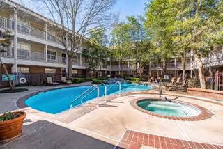 Outdoor view of a senior living facility courtyard featuring a swimming pool and a hot tub surrounded by a concrete deck. The area is enclosed by a three-story building with balconies and trees providing shade. Several lounge chairs are placed around the pool area under a clear blue sky.