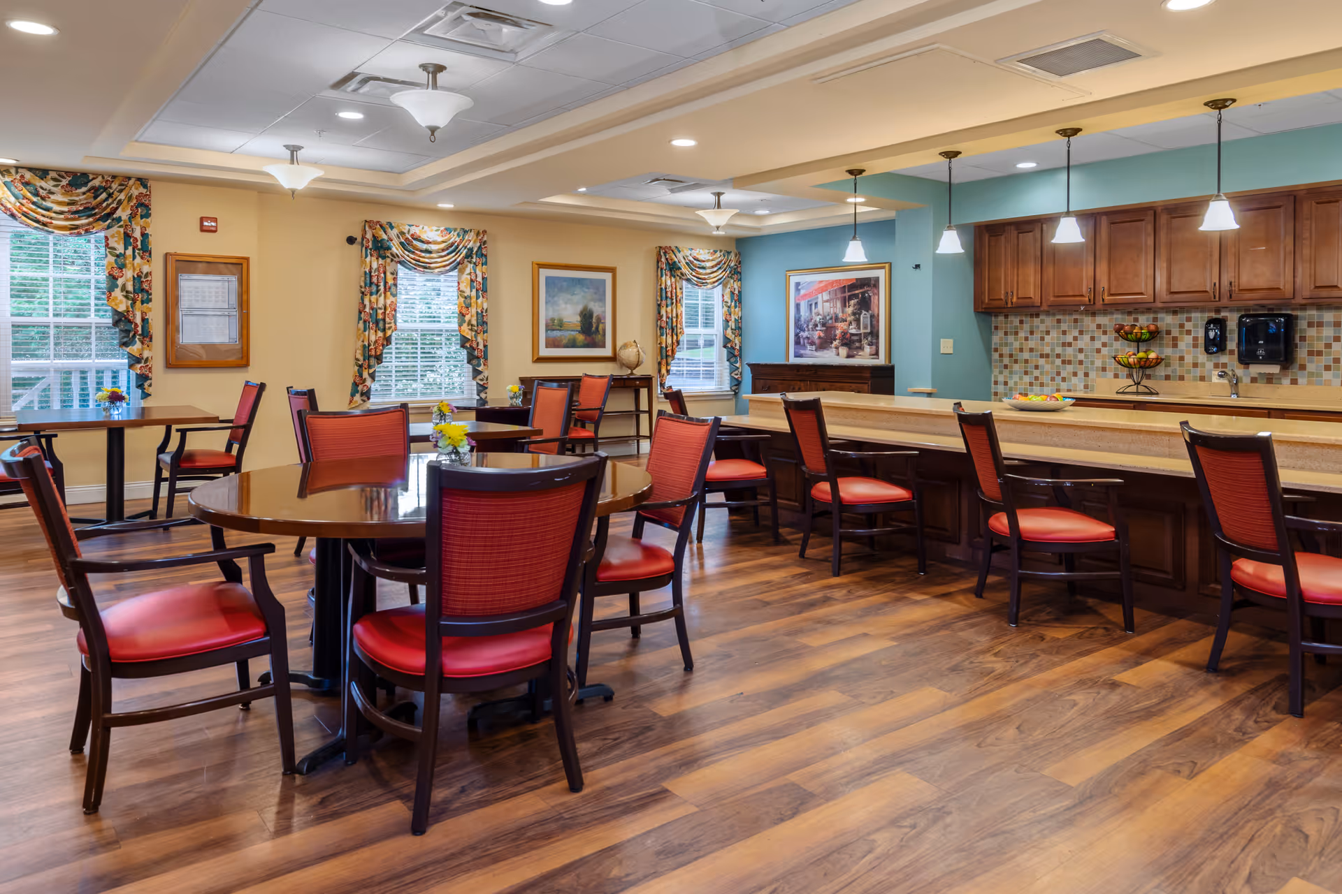 A bright dining room with several round and rectangular wooden tables surrounded by red cushioned chairs. The room features large windows with floral curtains, framed artwork on the walls, and a long counter with bar stools. The floor is wooden, and the ceiling has recessed lighting and hanging pendant lights above the counter.