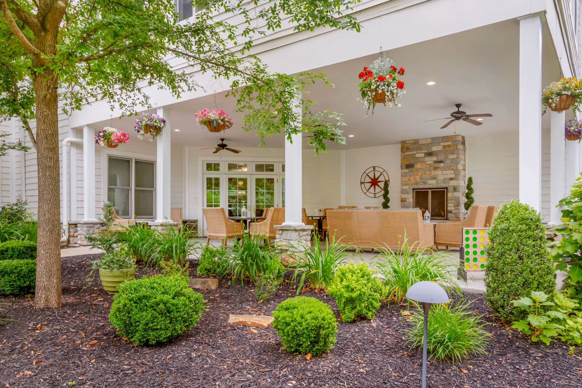 Covered outdoor patio area with wicker seating including chairs and sofas arranged around a stone fireplace. Hanging flower baskets are suspended from the ceiling, and ceiling fans are installed overhead. In the foreground, there is a landscaped garden with various green shrubs, plants, and a tree.