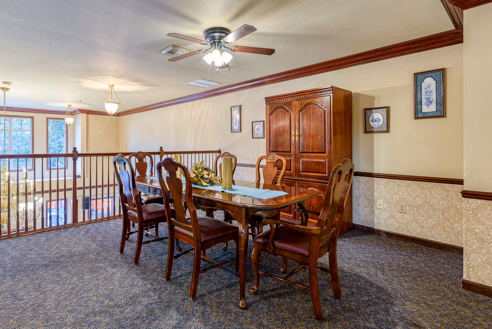 A dining area with a wooden dining table and six matching chairs. The table has a blue runner and decorative centerpiece. Behind the table is a wooden cabinet and three framed pictures on the wall. The room has beige walls with a patterned lower half, carpeted floor, and a ceiling fan with lights. The area overlooks a lower level with large windows and a chandelier.