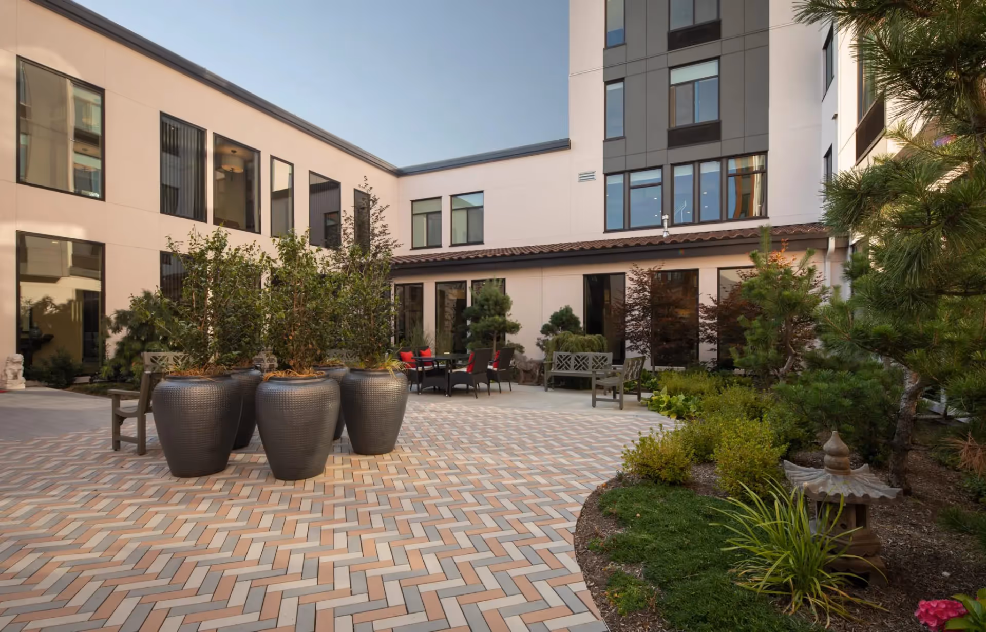 Outdoor courtyard area at Aegis Gardens Newcastle featuring large potted plants, benches, chairs with red cushions, and landscaped greenery surrounded by a multi-story building with large windows under a clear sky.