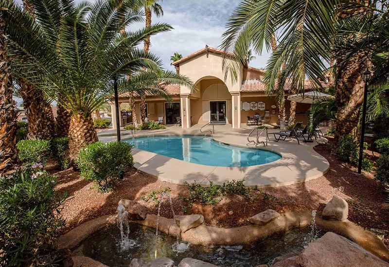 Outdoor view of a senior living facility courtyard featuring a kidney-shaped swimming pool surrounded by palm trees and landscaping. There is a small water fountain in the foreground with rocks and flowing water. The building in the background has a covered patio area with tables and chairs.