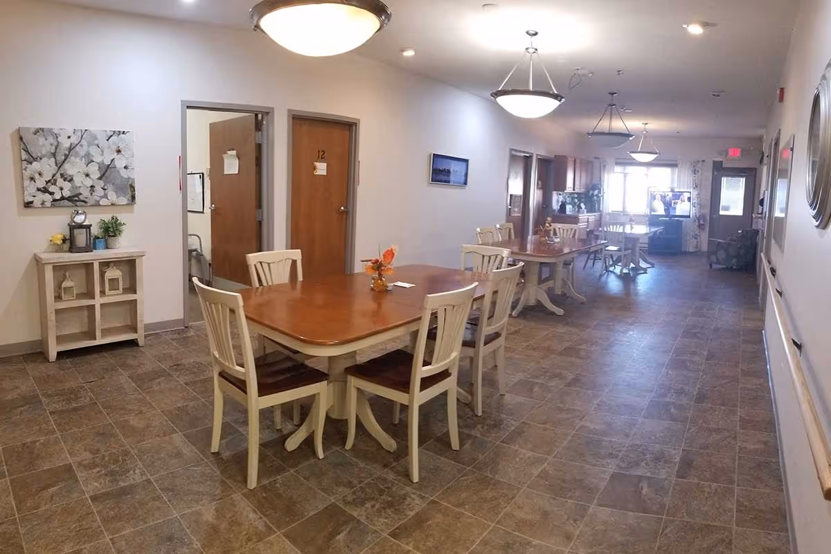 Interior view of a senior living facility dining area with multiple wooden tables and chairs arranged along a tiled floor. The room is well-lit with ceiling lights and has doors along the left wall, one marked with the number 12. There is a small cabinet with decorative items and a floral painting on the left wall. At the far end, a television is visible near a window with curtains.