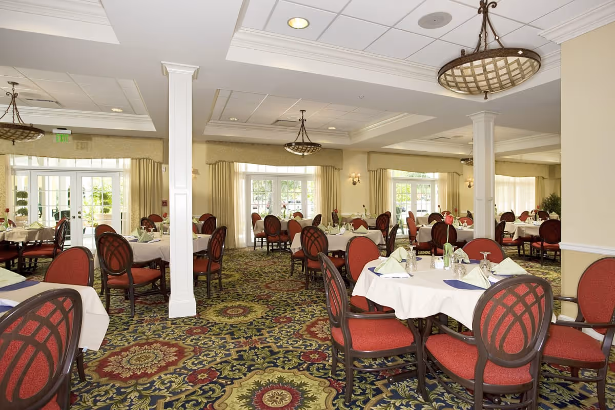 Spacious dining room with round tables set for meals, red upholstered chairs, chandeliers, and a patterned carpet.