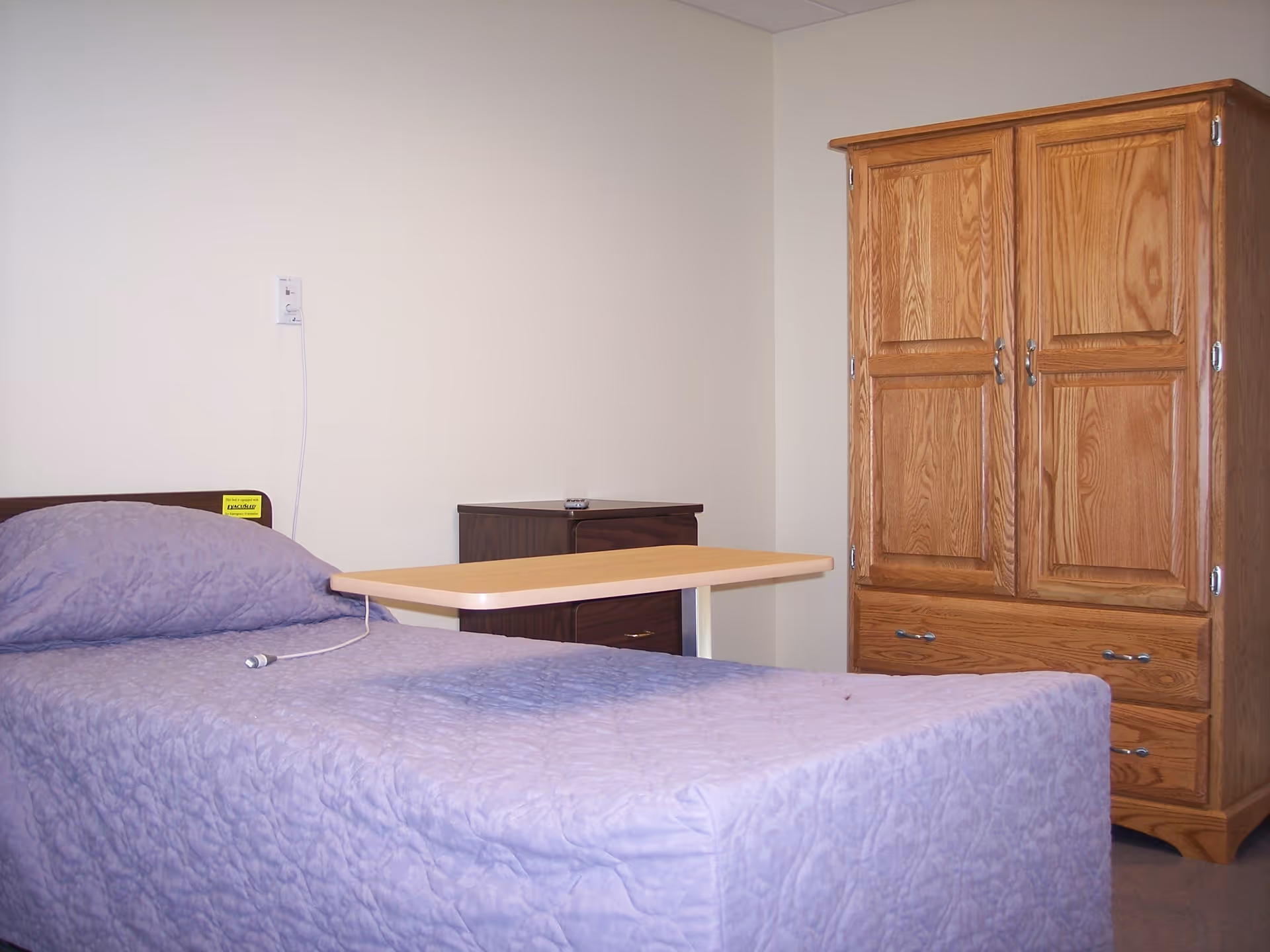 A simple bedroom with a single bed covered in a light purple quilt, a wooden bedside table with a small item on top, a wooden wardrobe with two doors and two drawers, and a beige wall with a call button and cord hanging beside the bed.