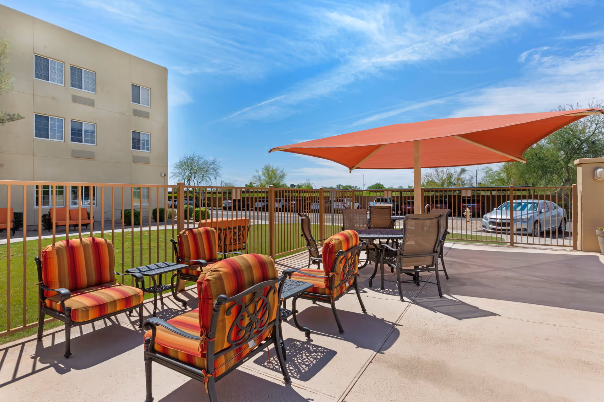 Outdoor patio area with striped cushioned chairs and metal tables under a large red umbrella, adjacent to a beige building with multiple windows, and a parking area with cars in the background under a blue sky.