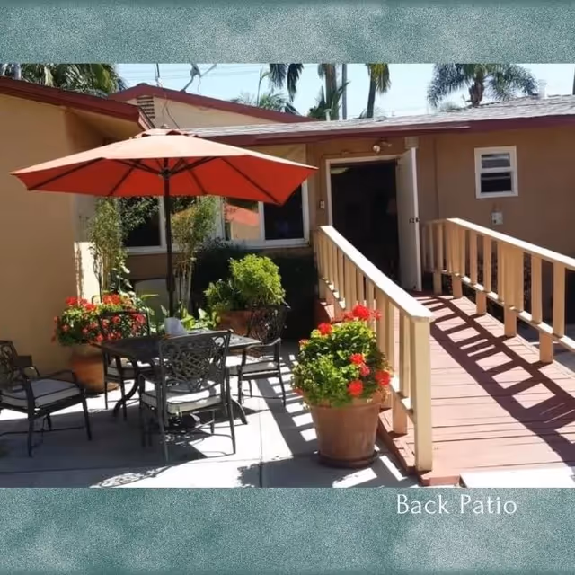 Back patio with a dining table and chairs under a red umbrella, potted flowers, and a wooden ramp leading to a doorway.