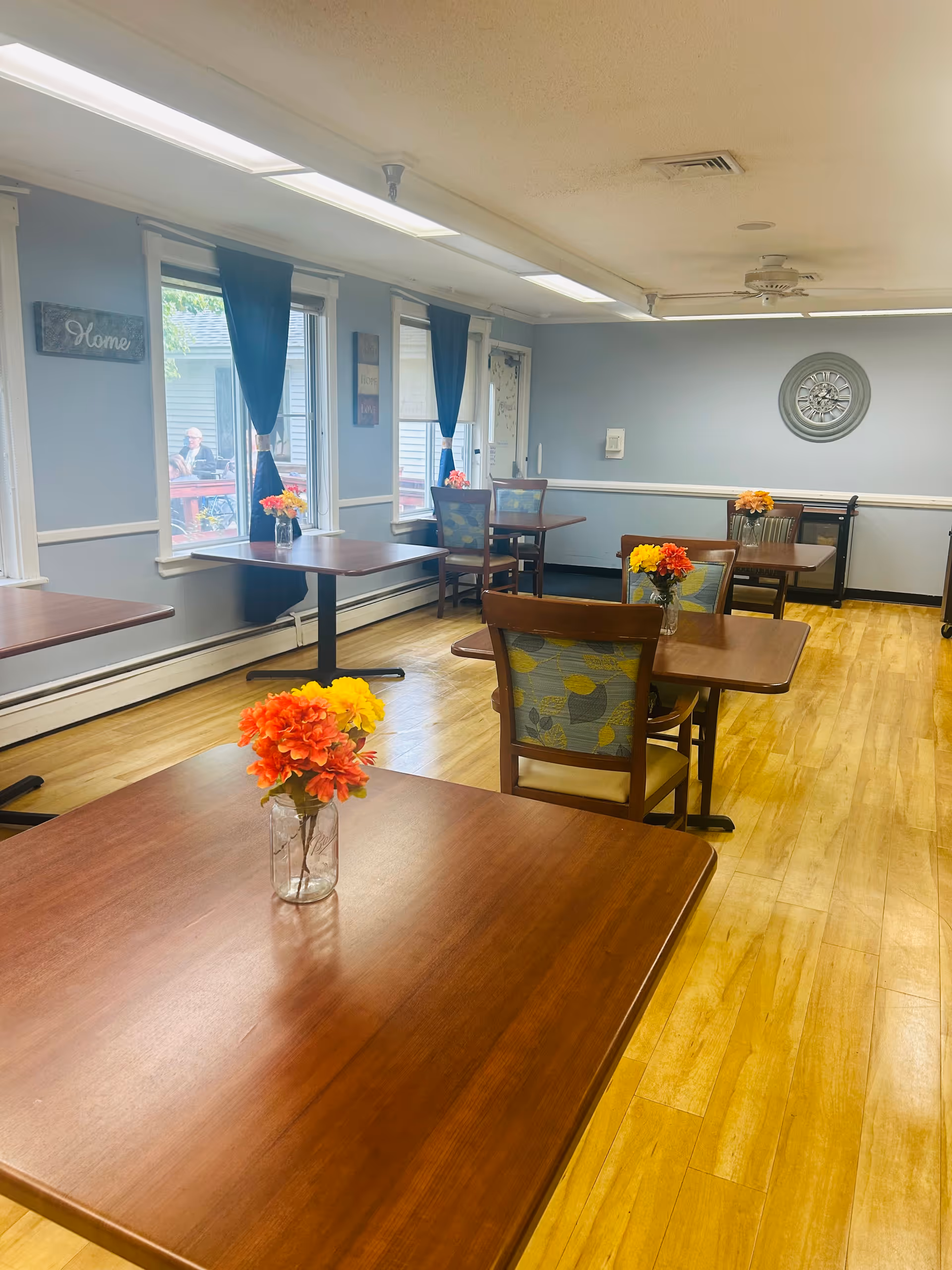 A dining room with wooden tables and chairs, each table decorated with a small vase of colorful flowers. The room has light blue walls, large windows with dark curtains, and wooden flooring. There is a decorative wall clock and a 'Home' sign on the wall.