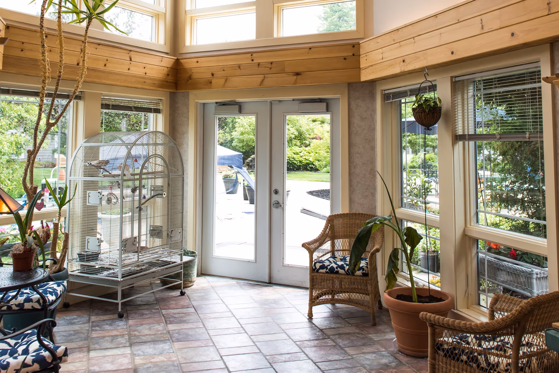 A bright sunroom with large windows and glass double doors leading outside. The room features tiled flooring, wicker chairs with patterned cushions, several potted plants, and a large birdcage. Natural light fills the space, highlighting the wooden trim and greenery visible outside.