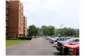 Parking lot with multiple parked cars alongside a multi-story brick building on the left and trees in the background under a cloudy sky.