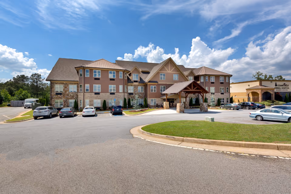 Exterior view of a senior living facility named Oaks at Acworth, showing a large multi-story building with a covered entrance, several parked cars, and a clear blue sky with clouds.
