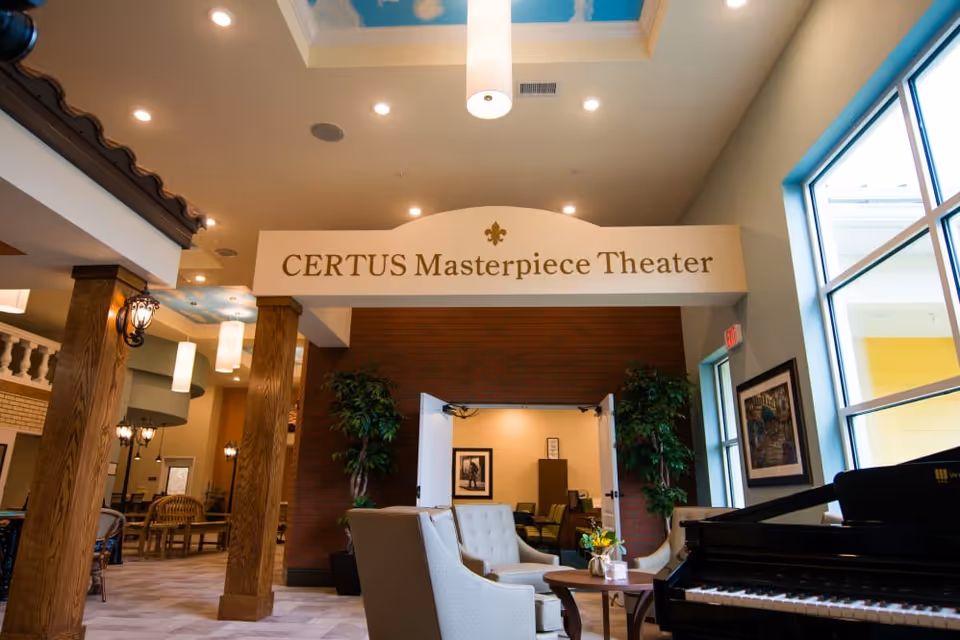 Interior view of CERTUS Premier Memory Care Living showing the entrance to the CERTUS Masterpiece Theater. The area features comfortable seating with armchairs and a small table, a grand piano, large windows letting in natural light, wooden pillars, and decorative plants. The ceiling has a light fixture and a painted sky design.
