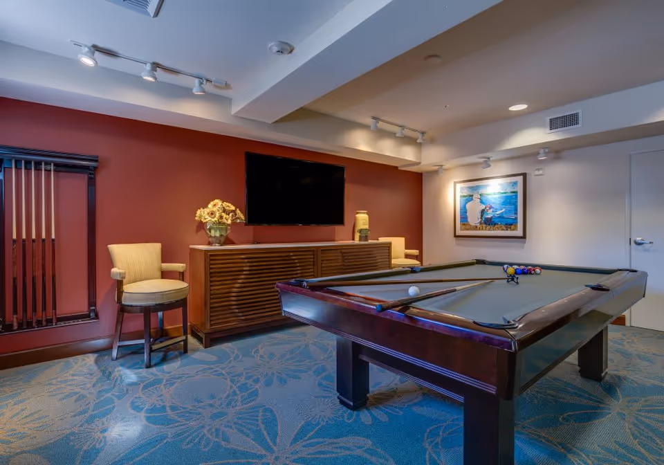 A recreational room with a pool table in the center, two beige chairs, a wooden cabinet with a vase of flowers and a decorative item, a wall-mounted flat screen TV, and a framed painting on the wall. The room has a red accent wall and blue carpet with floral patterns.