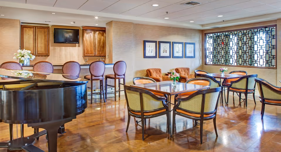 Communal dining and lounge area with a grand piano, multiple tables and chairs, bar seating and a decorative partition window.