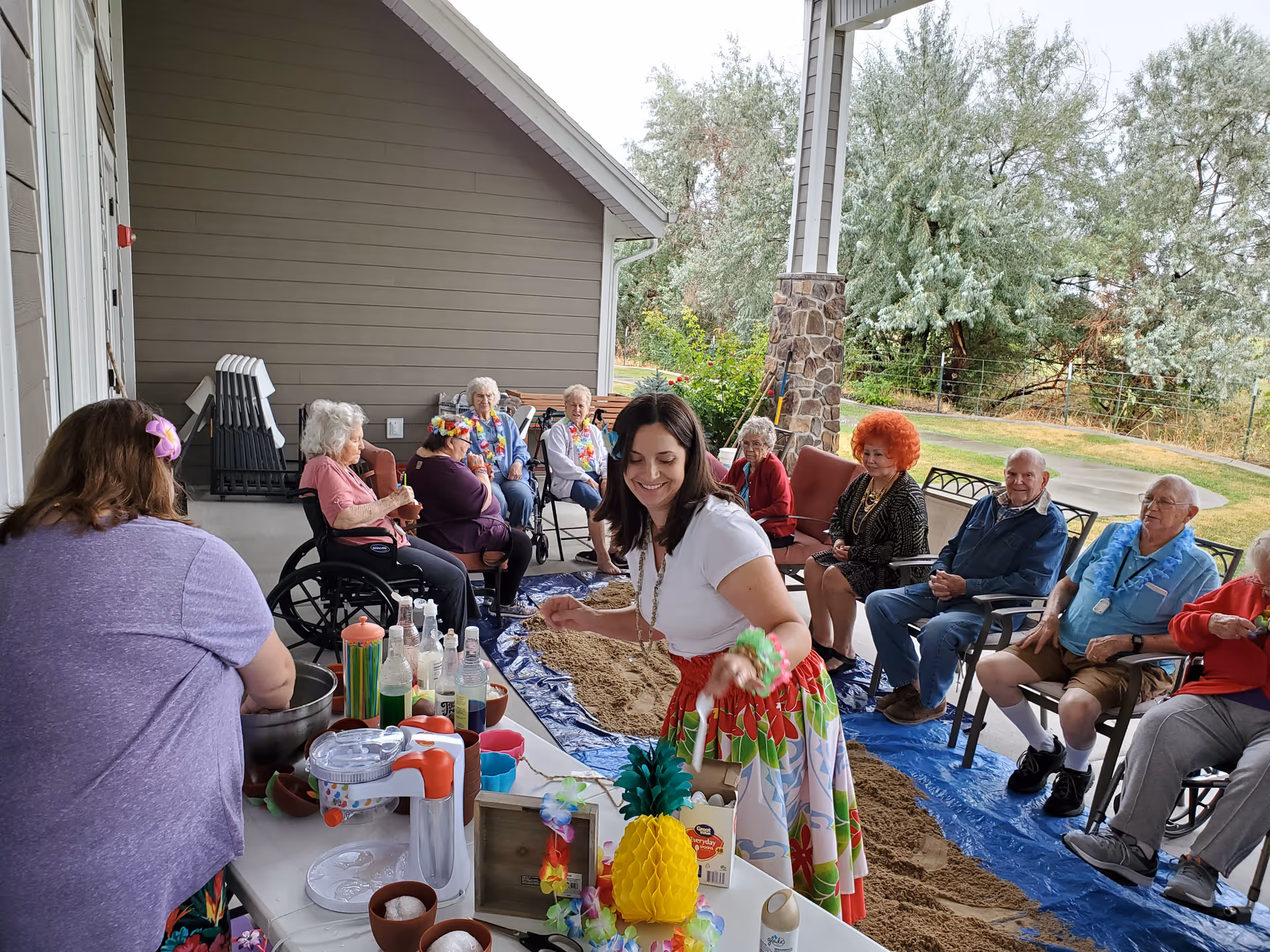 A group of elderly people sitting in chairs and wheelchairs on a covered patio area, participating in a social event with a tropical theme. A woman in a colorful skirt and white top is serving drinks or snacks at a table decorated with tropical items like a pineapple and leis. The patio overlooks a grassy outdoor area with trees.