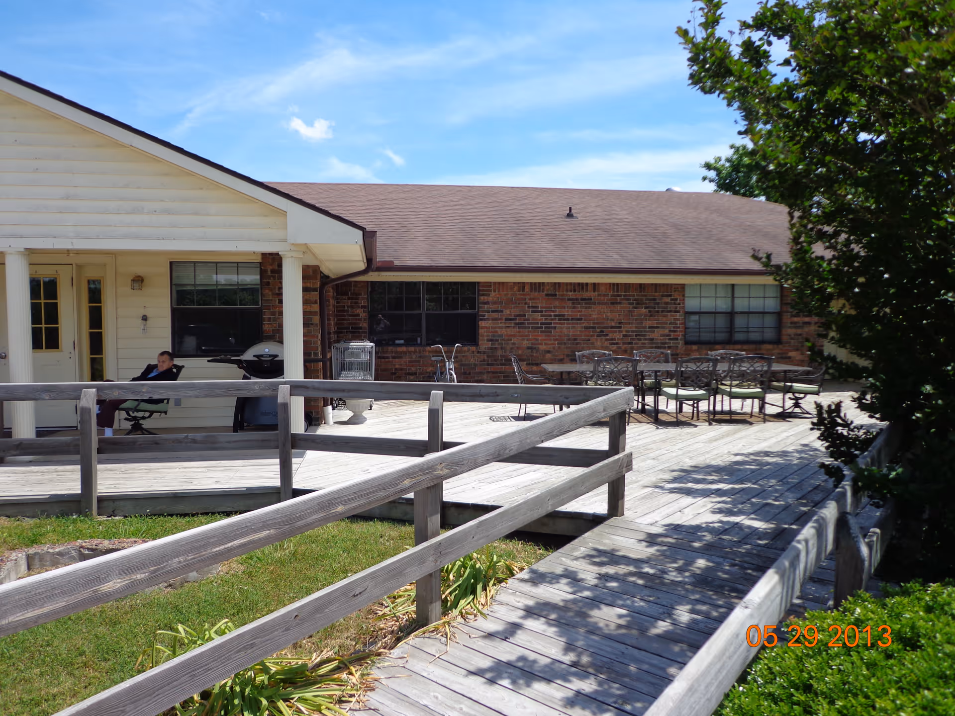 Outdoor wooden deck area attached to a single-story building with brick and white siding. The deck has a wooden railing and a ramp leading up to it. There is a person sitting on a chair near a grill on the left side of the deck. On the right side, there is a table with several chairs. Trees and greenery surround the deck under a clear blue sky.