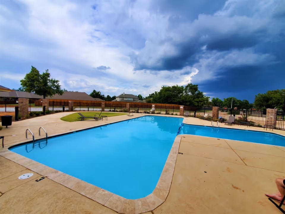 Outdoor swimming pool with clear blue water surrounded by a concrete deck and a few lounge chairs. The pool area is enclosed by a black metal fence with brick pillars. Trees and houses are visible in the background under a partly cloudy sky with dark storm clouds approaching.