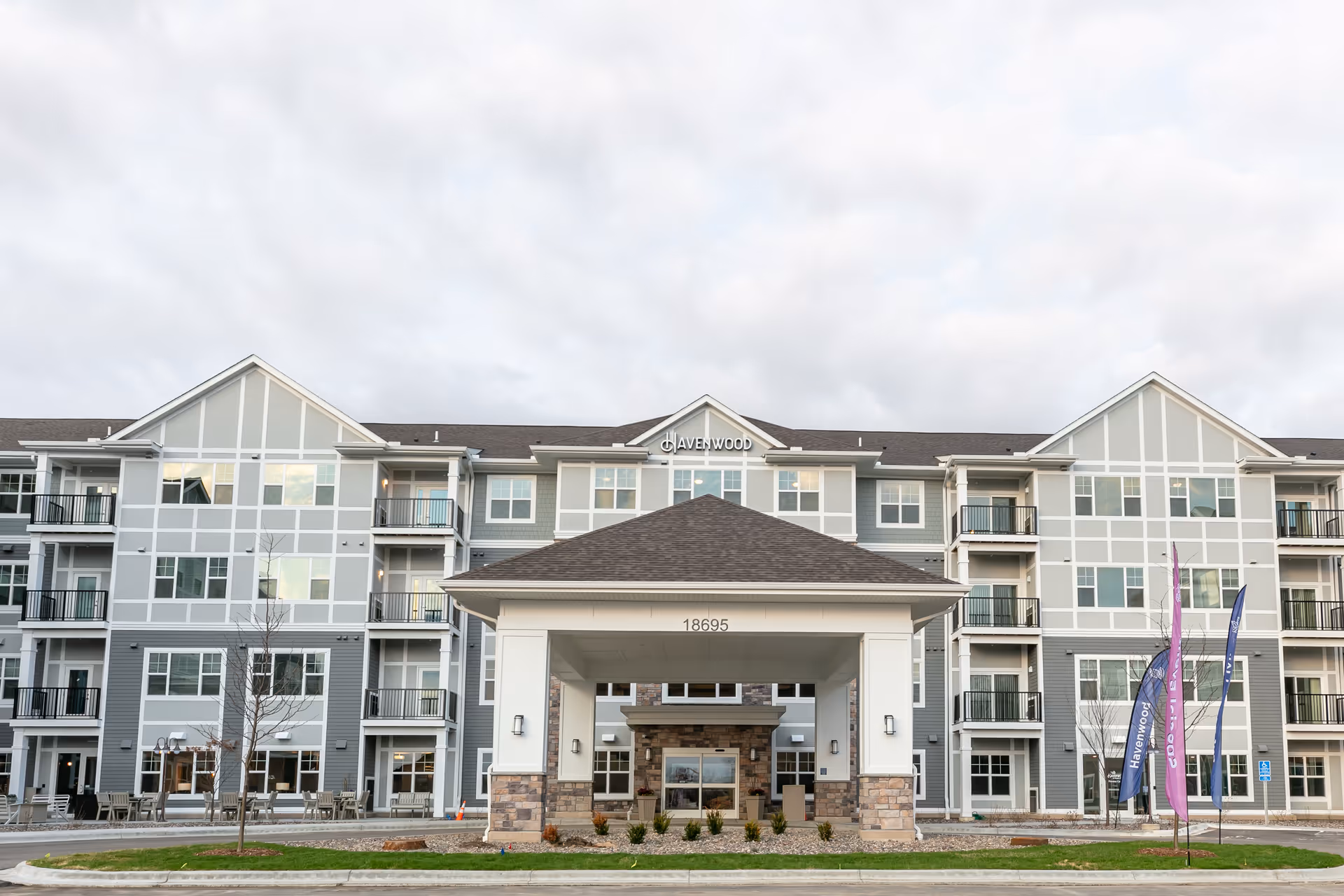 Front exterior view of Havenwood of Maple Grove, a multi-story senior living facility with a covered entrance, balconies, and large windows. The building is gray with white trim and has flags on the right side near the entrance.