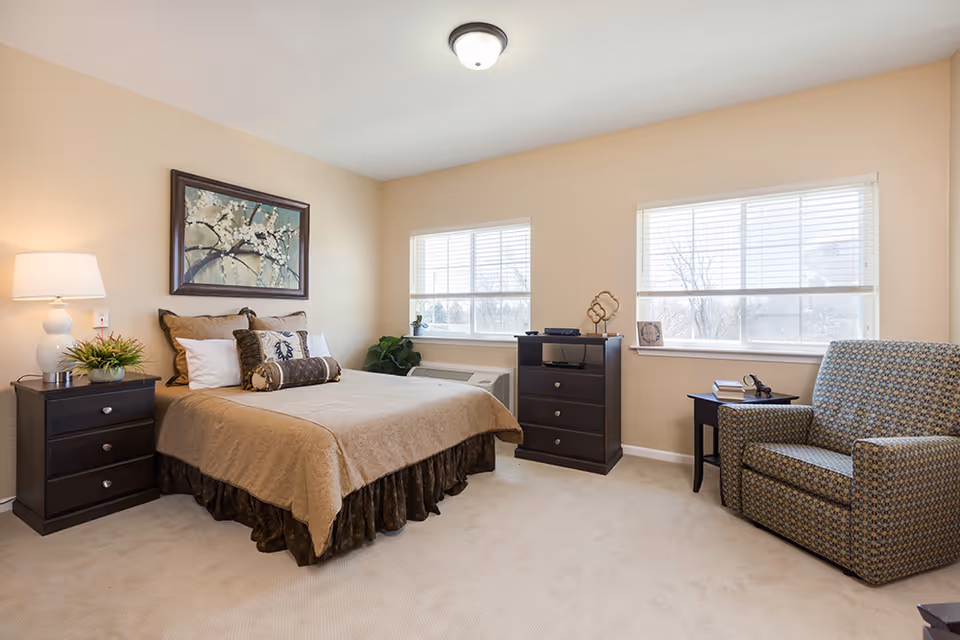 A well-lit bedroom in an assisted living facility featuring a neatly made bed with beige and brown bedding, two windows with blinds, a dark wooden nightstand with a lamp and plant, a matching dresser with decorative items, and a patterned armchair next to a small side table with books and glasses.