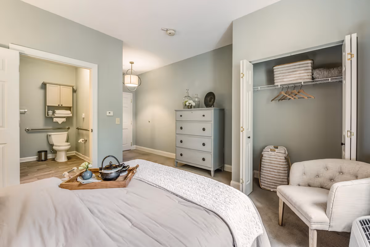 A bedroom with a bed in the foreground, a wooden tray with a teapot and cup on the bed, an open closet with hangers and storage baskets, a beige upholstered chair, a gray chest of drawers with decorative items on top, and an open door leading to a bathroom with a toilet and wall-mounted cabinet.