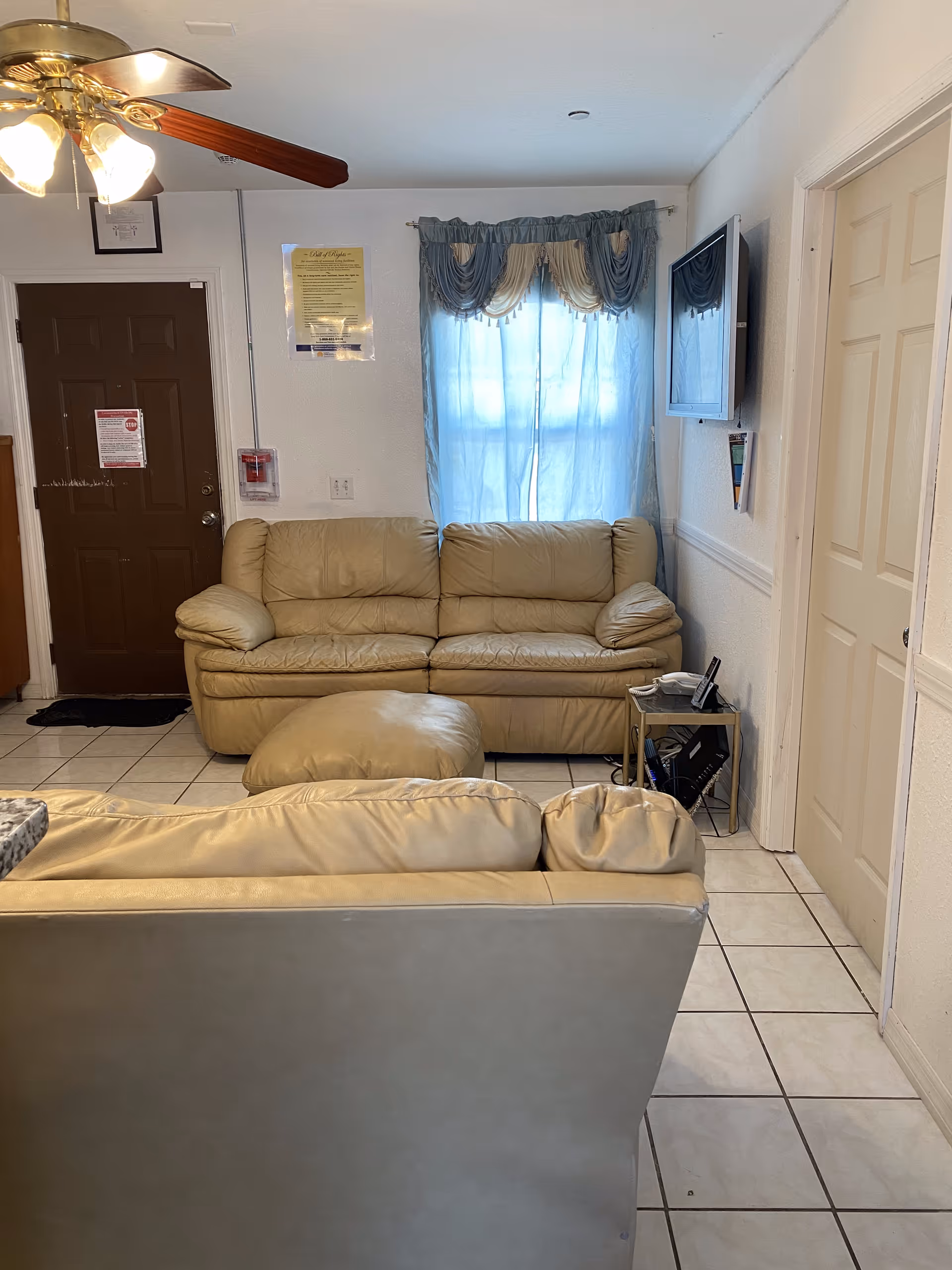 A small living room with beige leather sofas and an ottoman. There is a window with blue and cream curtains behind one sofa. A flat-screen TV is mounted on the wall to the right. A ceiling fan with lights is visible on the ceiling. The floor is tiled with white tiles, and there is a brown door on the left side of the room.