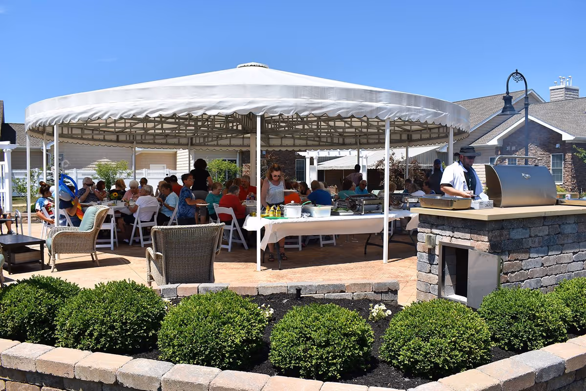 Outdoor patio area at The GreenFields Continuing Care Community with a large white canopy covering several tables where people are seated and enjoying a meal. There is a stone grill station with a person preparing food, surrounded by neatly trimmed bushes and a clear blue sky.