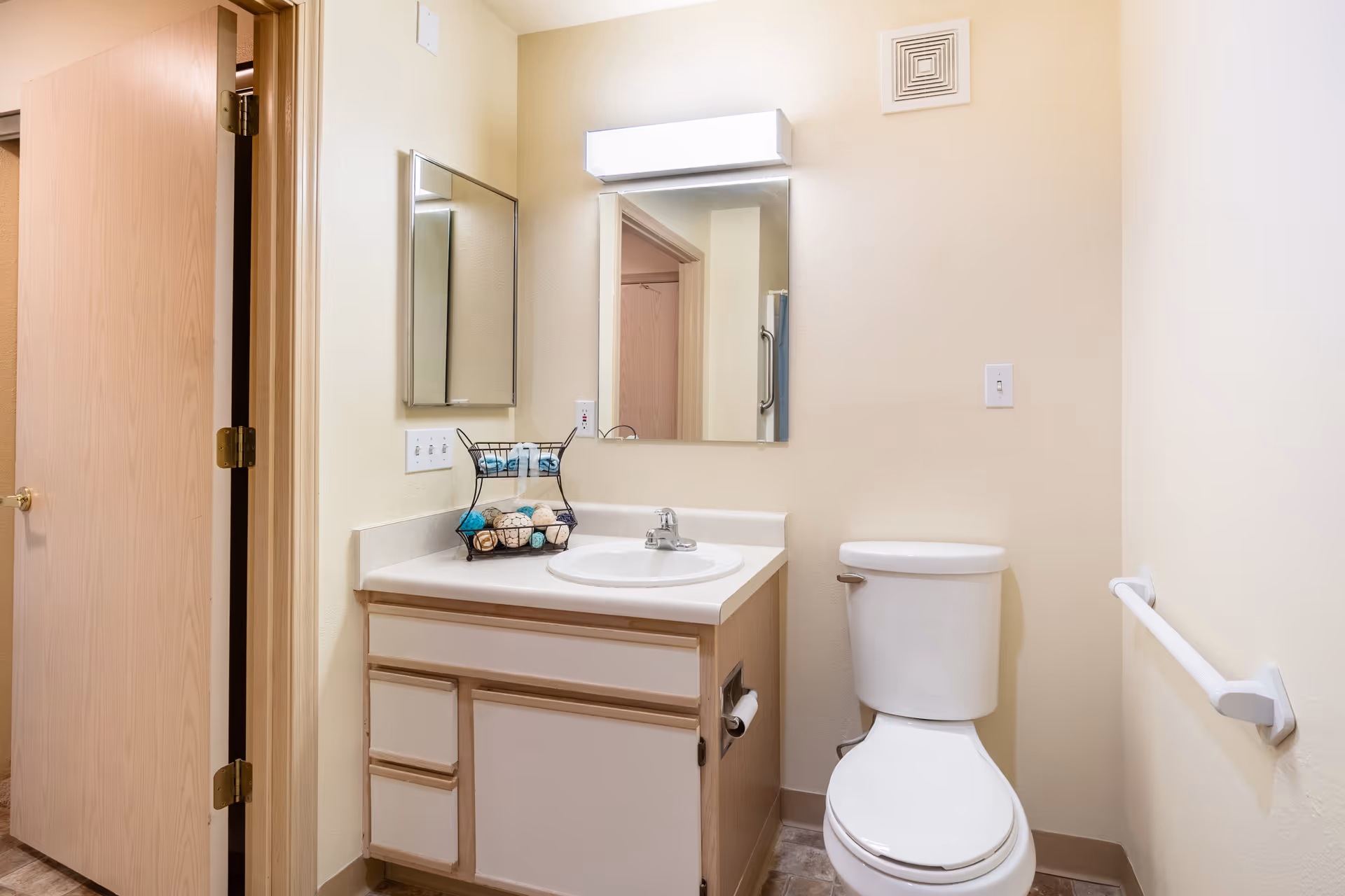 A clean and simple bathroom with a white toilet, a vanity with a sink, a mirror above the sink, and a small decorative basket with rolled towels on the countertop. The walls are light-colored, and there is a grab bar next to the toilet. A wooden door is partially open, revealing another room.