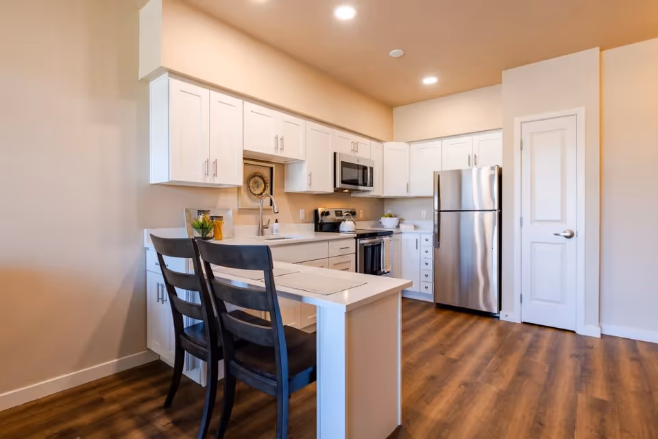 Modern kitchen with white cabinets, stainless steel refrigerator, stove, and microwave. There is a small island with two dark wooden chairs and wood flooring throughout.