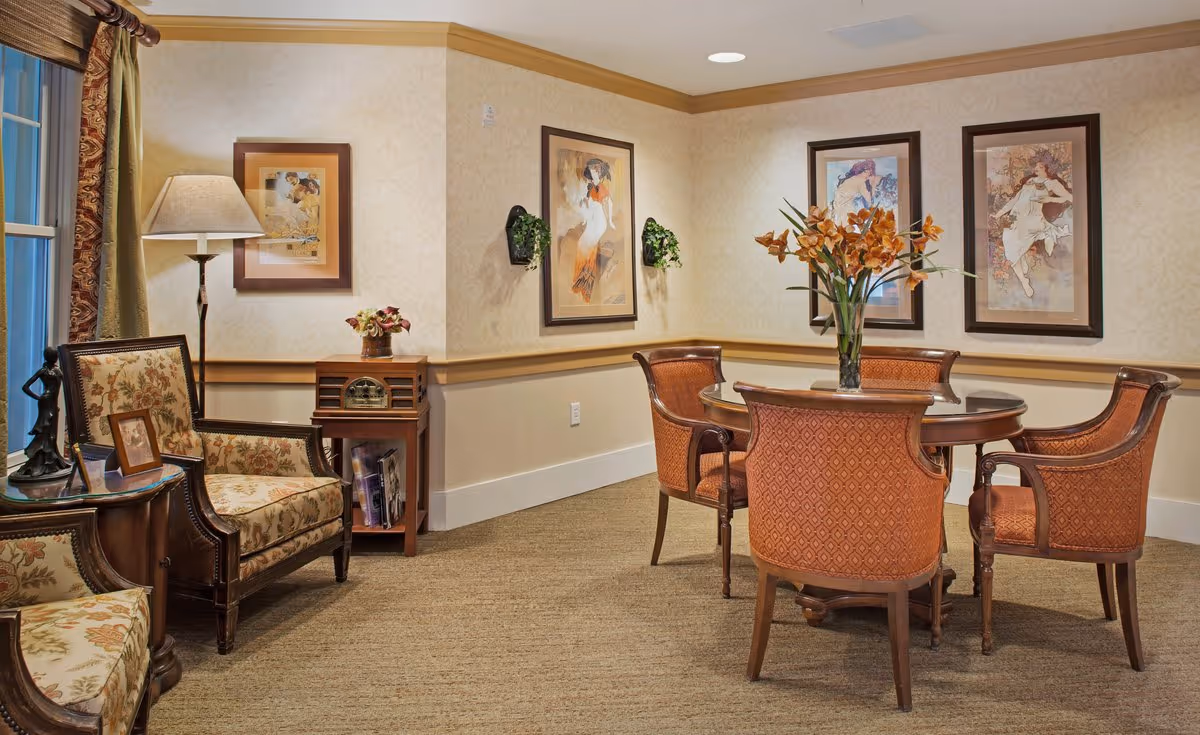 Cozy senior living common room with upholstered armchairs, a round wooden table with four chairs, framed artwork, and a floor lamp.