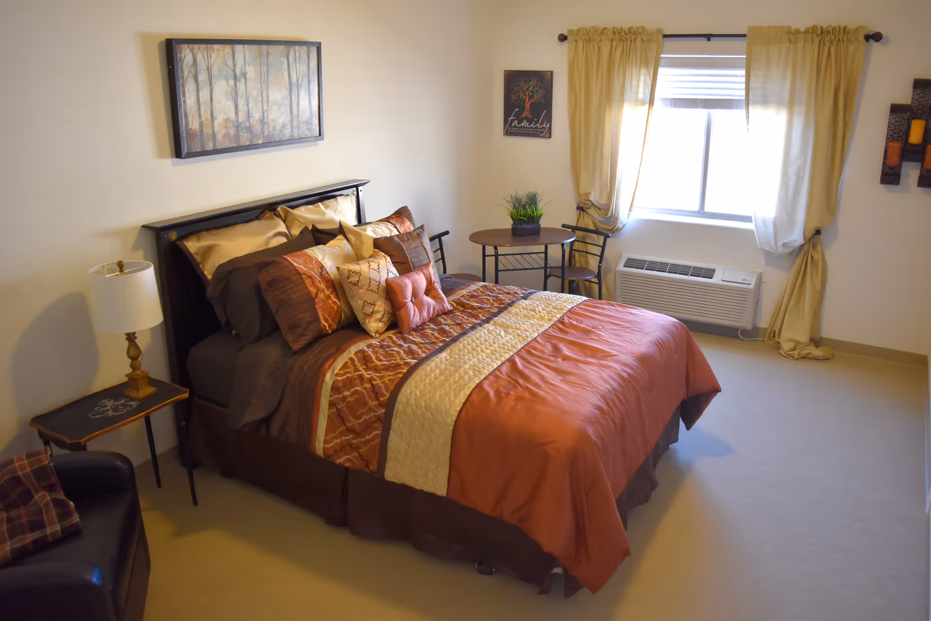 A cozy bedroom with a neatly made bed featuring brown, orange, and cream bedding. There is a small side table with a lamp next to the bed, a black armchair with a plaid pillow, a round table with two chairs near a window with beige curtains, and wall decorations including a framed picture above the bed and a small 'family' sign on the wall.