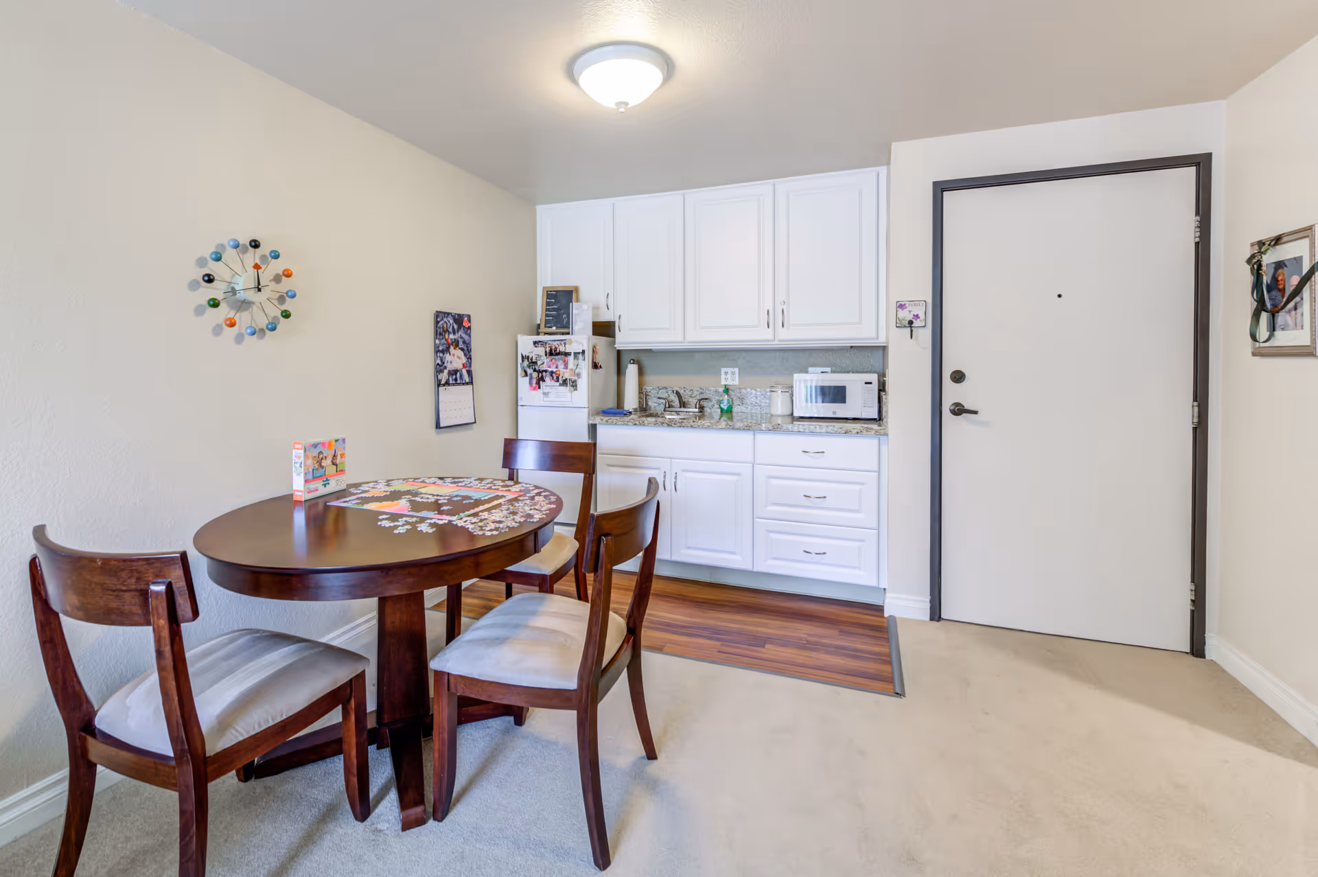 A small dining area with a round wooden table and three chairs with cushioned seats. On the table, there is a partially completed jigsaw puzzle. Behind the table is a compact kitchenette with white cabinets, a small refrigerator, a microwave, and a sink. A colorful wall clock and a calendar are mounted on the wall. To the right, there is a white door with a dark frame and a picture hanging on the adjacent wall.