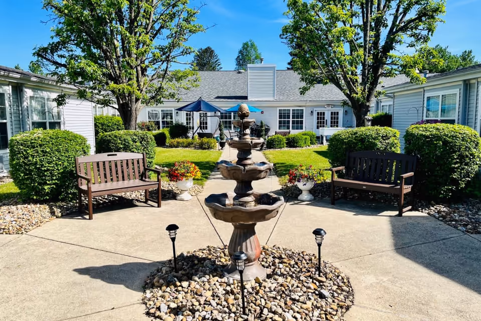Sunny courtyard with a central multi-tiered fountain, benches, potted flowers, trimmed bushes, and surrounding single-story buildings.