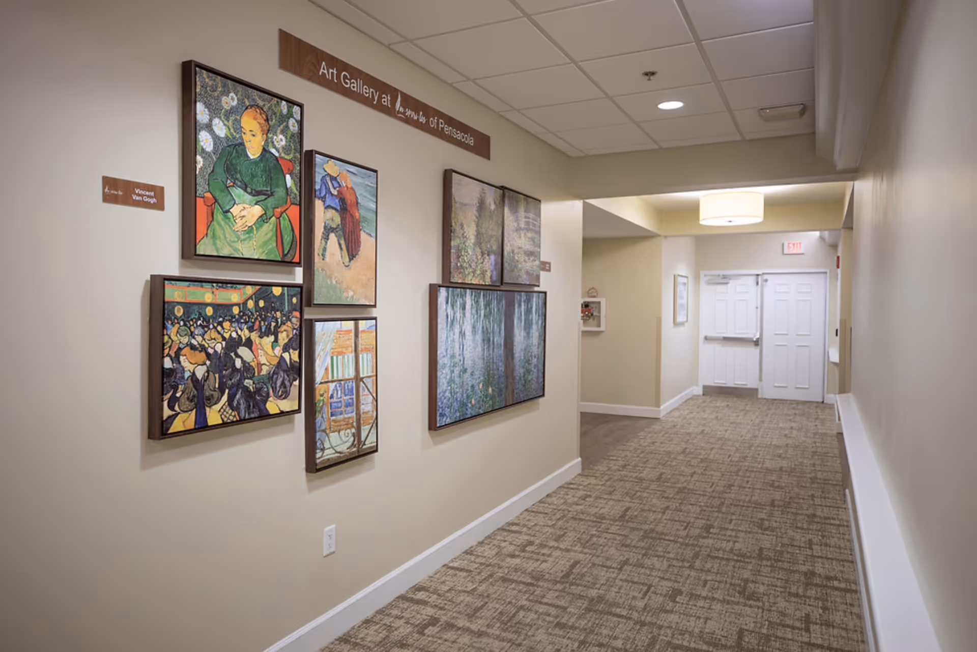 A hallway in a senior living facility with beige walls and carpeted floor. On the left wall, there is an art gallery display featuring framed paintings, including works inspired by Vincent Van Gogh. The hallway is well-lit with ceiling lights and leads to a set of double doors at the end.