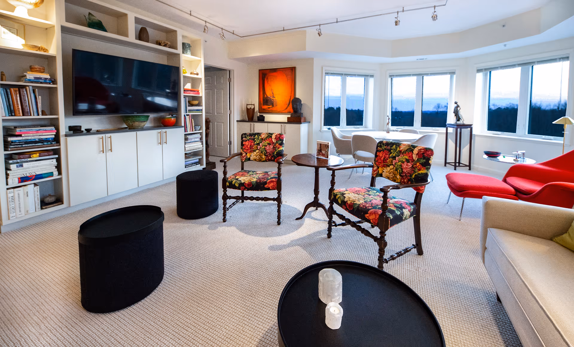 A bright living room with large windows showing an outdoor view. The room features a beige carpet, a built-in white shelving unit with a flat-screen TV, two floral upholstered wooden chairs around a small round wooden table, a beige sofa with a green cushion, a red modern armchair with an ottoman, and two black round ottomans. There are various decorative items on the shelves and tables, and a piece of artwork on the wall near the door.