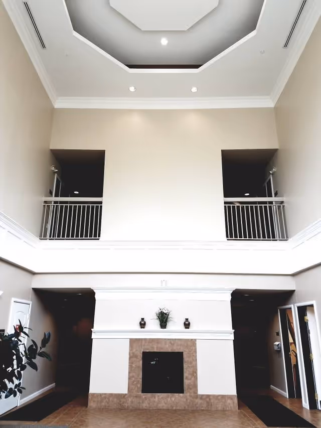 Interior view of a spacious room with a high ceiling and a decorative recessed ceiling light. The room features a fireplace with a tiled surround and mantle decorated with two small vases and a plant. There are two open balcony areas with white railings above the fireplace, and doorways on either side of the fireplace leading to other rooms. A plant is visible on the left side near the doorway.